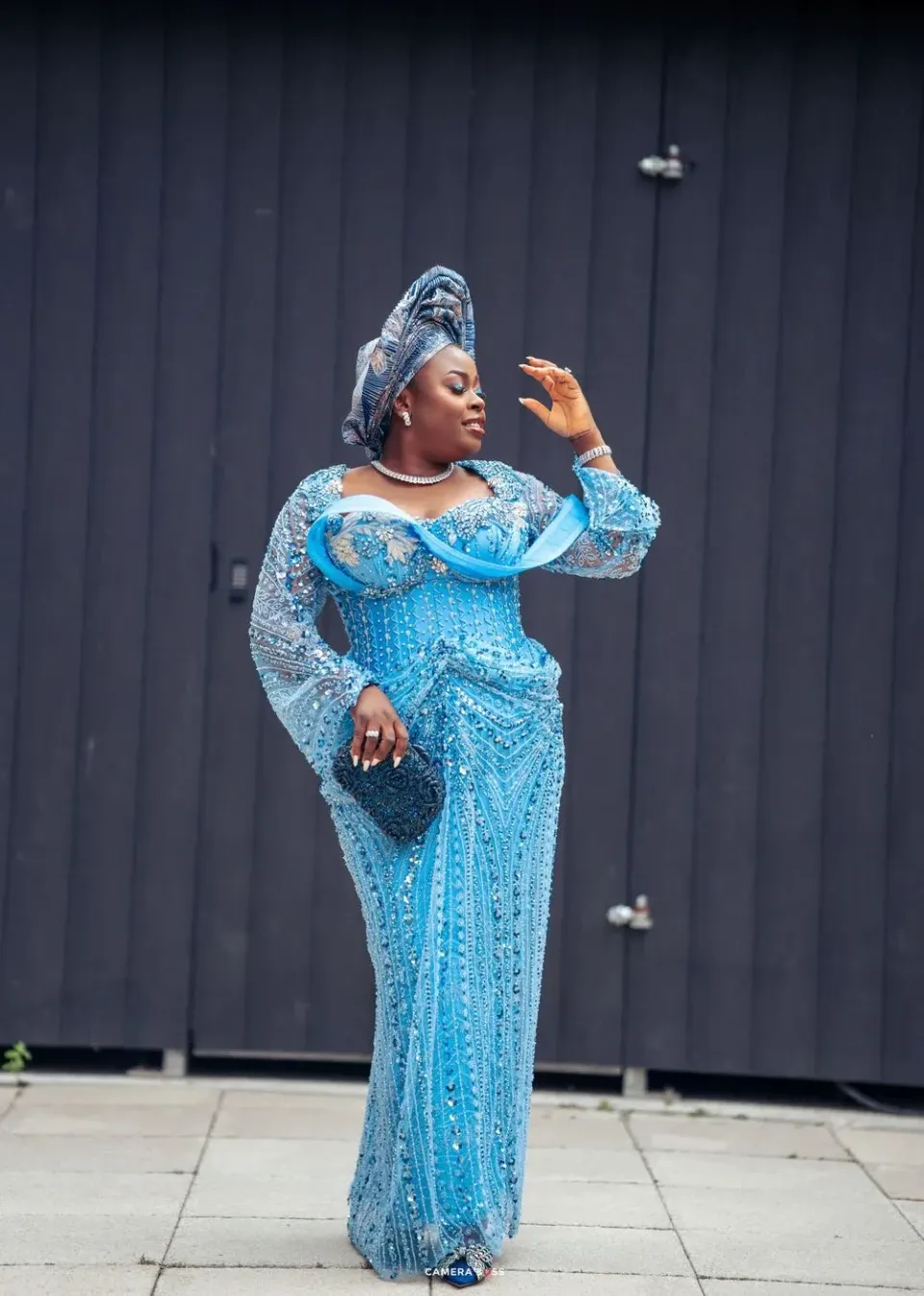 A person wearing an elegant turquoise blue sequined gown and matching head wrap poses dramatically against a dark wall.