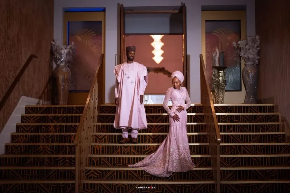 Two people in elegant pink formal attire pose on a grand staircase in an ornate interior setting.