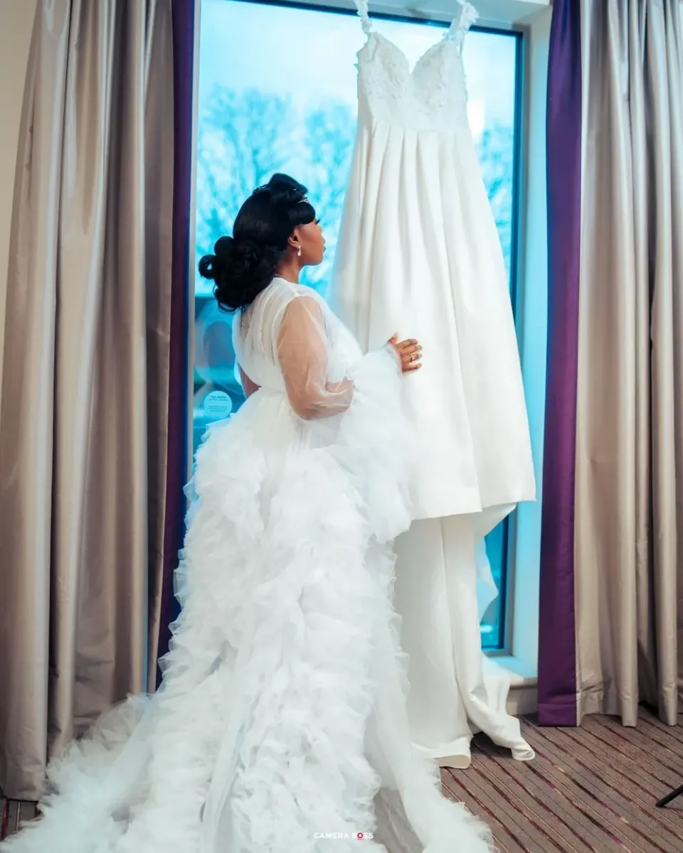 A figure in a white ruffled gown stands looking at a classic white wedding dress displayed by a window.